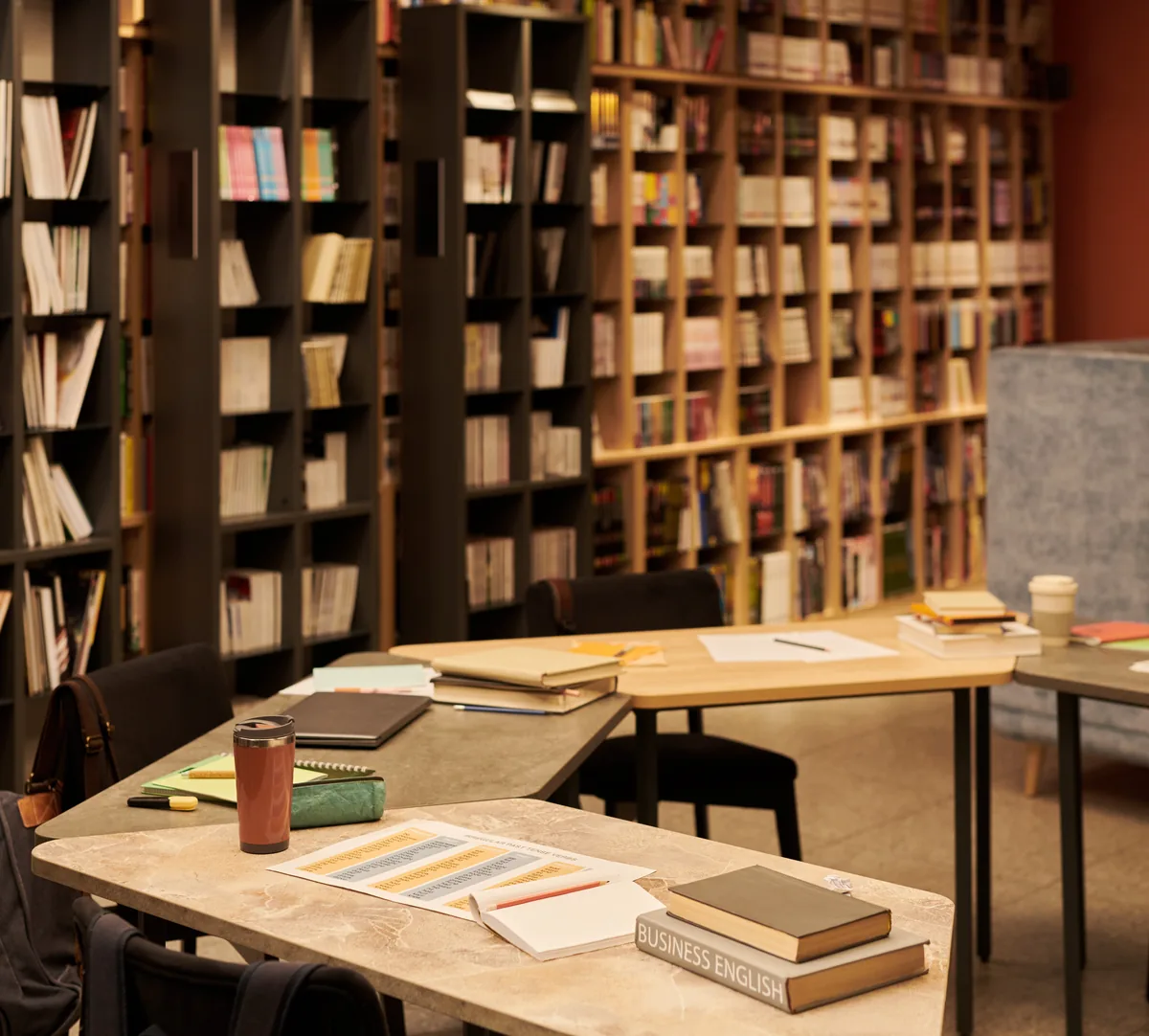 A library reading room: open books, notes and study materials spread across a desk in front of full shelves